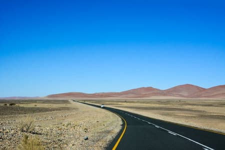 A long asphalt road in the desert in perspective against a blue sky. A vehicle drives in the distance along the road. mountains in the background