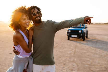 Image of happy african american couple pointing finger and walking while travelling with car on desert